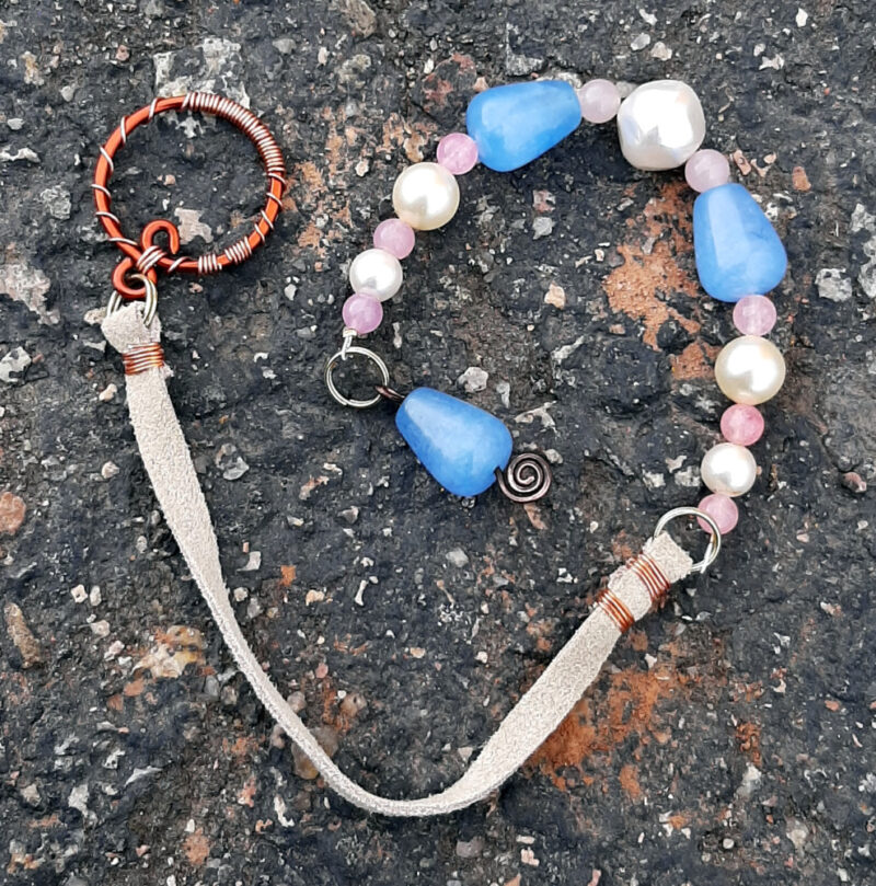 A pendulum; the bottom is a vaguely cone-shaped pale blue bead threaded onto silver-colored wire bent into a spiral shape at the bottom. Above that are alternating pink and white beads, interspersed with two more of the blue ones. A length of narrow white leather leads to a round loop of bright copper wire decoratively wrapped in much narrower copper wire.