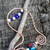 A driftwood stick hangs down, angled to the left; wired to it is a long arc in hammered copper wire, with a spiral on each end. Between the spirals are a large glass bead patterned in red, blue, white, and teal, with smaller white beads to either side. Crossing the top of the arc, above the driftwood, is a draped arch of alternating blue and white beads.