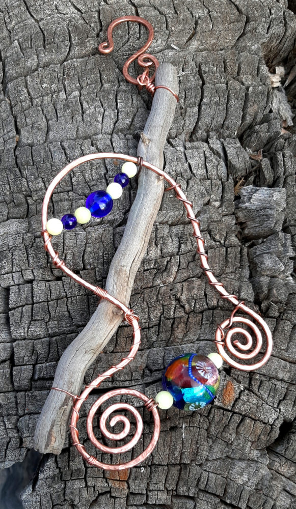 A driftwood stick hangs down, angled to the left; wired to it is a long arc in hammered copper wire, with a spiral on each end. Between the spirals are a large glass bead patterned in red, blue, white, and teal, with smaller white beads to either side. Crossing the top of the arc, above the driftwood, is a draped arch of alternating blue and white beads.