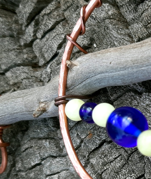 The spot where the copper arc meets the driftwood. Thinner copper wire wraps around the thick stuff, ducking under the stick to hold the two together before heading off, bearing blue and white beads.