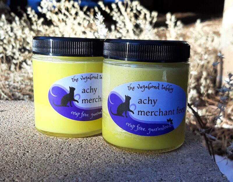 Two big clear glass jars filled with pale yellow salve. The labels say 'achy merchant feet'.
