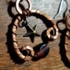 Broad arches of hammered copper wire, the ends of each curved inward with a maroon oval glass bead suspended between. Small brass stars hang in the center.