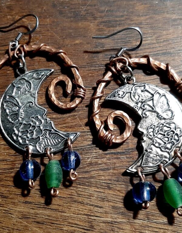 Hammered copper arches, one end spiraling inward while the other end curves out to a simple loop. Inside hang crescent moons, but broad, almost half moons, with butterflies and leaves embossed inside. From the bottom of each moon hang beads, blue green blue, each from its own loop.
