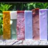 A stack of six bars of soap in various colors -- yellow, green, red, pale blue, variegated brown, and purple.