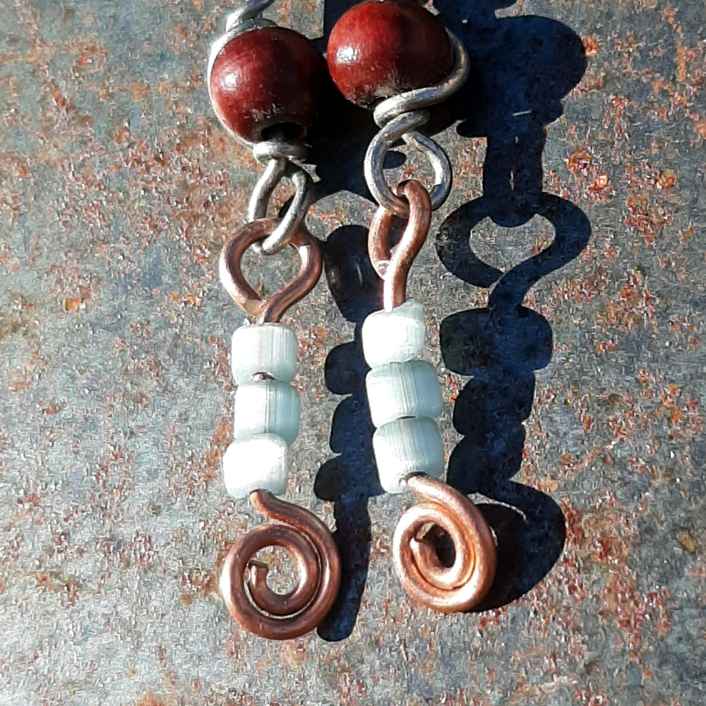 Each earring has two sections. The lower has three pale blue beads strung on copper wire with a neat spiral at the bottom. This is linked to a piece with a round red wooden bead strung on steel wire that curves back up around the bead, then twists around itself.