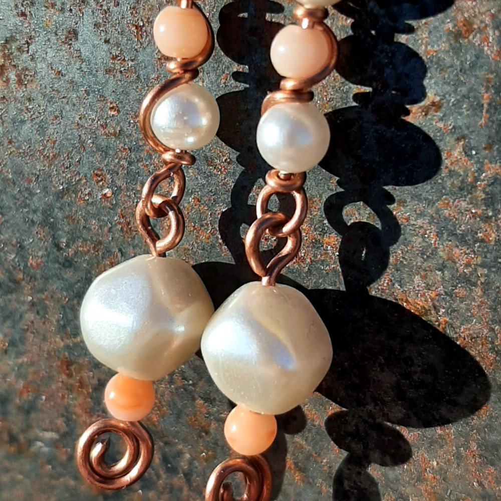 Each earring has two sections. The lower has a round white bead with a much smaller peach bead below it, both strung on copper wire with a neat spiral at the bottom. This is linked to a piece with another peach bead between two white beads, all strung on copper wire that curves back up around the beads, then twists around itself.