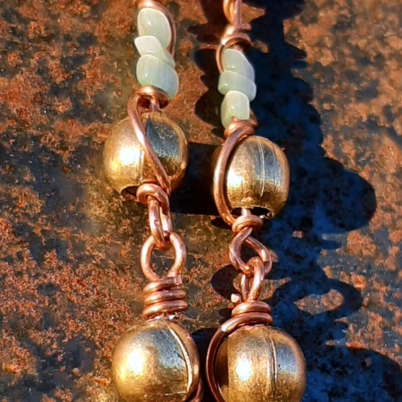 Each earring has two sections. The lower has a round brass bead strung on copper wire with a neat loop at the bottom. This is linked to a piece with another brass bead with three pale blue beads above it, all strung on copper wire that curves back up around the beads, then twists around itself.