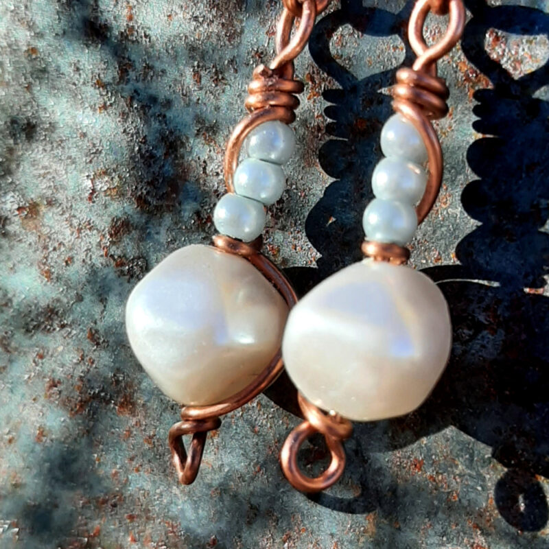 Copper wire earrings, hinged at the middle; at the top, copper wire wrapping around itself; below, a single white bead with three tiny pale blue beads above.