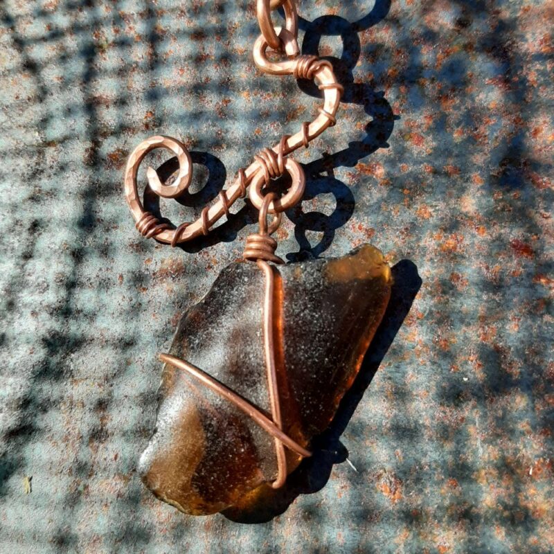 A coke-bottle brown piece of creek glass, wrapped in copper wire, hangs from a curled and curved piece of much thicker hammered copper wire.