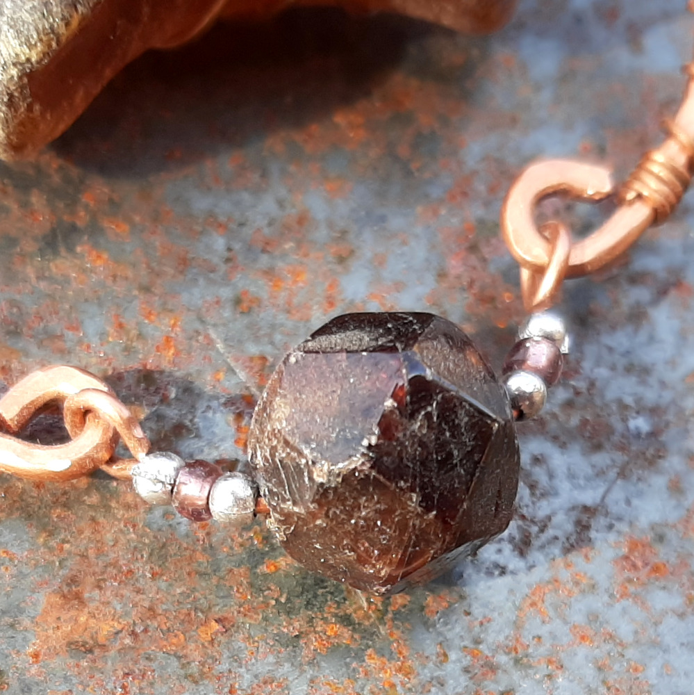 This piece of brown creek glass would be called a cylinder if it weren't also arched. It's simply wrapped in copper wire, as is the near-circle of hammered copper wire around it. Below, in between the ends of the near-circle, hangs a roughly faceted hunk of garnet between a hew much smaller purple and silver-colored beads.