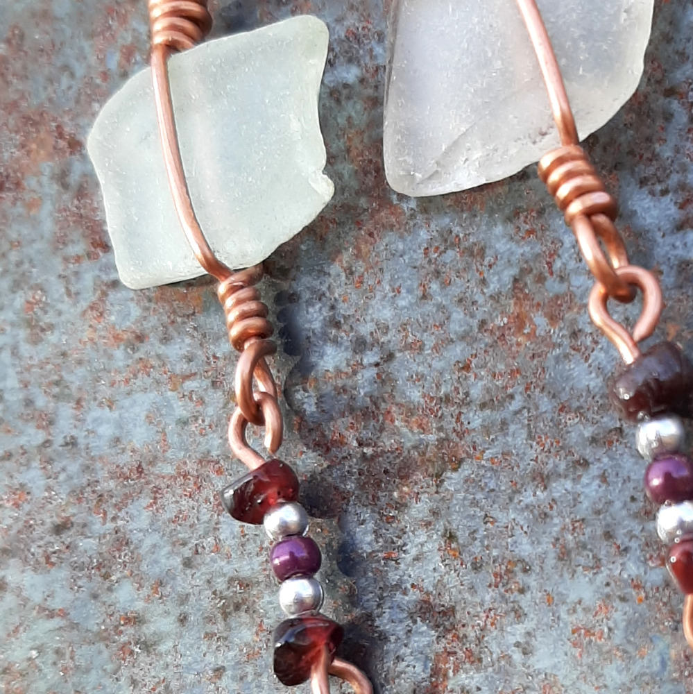 Each earring has a roughly rectangular piece of white creek glass, simply wrapped in copper wire, from which hangs another short length of copper with purple and silver-colored beads threaded onto it.