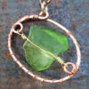 A very vaguely roundish piece of green creek glass, simply wrapped in gold-colored wire, sits entirely within an oval of hammered copper wire. The oval sits at an angle, one short end pointing slightly upward.