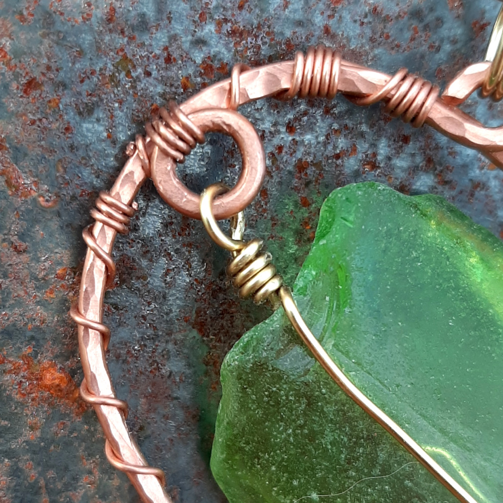 A very vaguely roundish piece of green creek glass, simply wrapped in gold-colored wire, sits entirely within an oval of hammered copper wire. The oval sits at an angle, one short end pointing slightly upward.