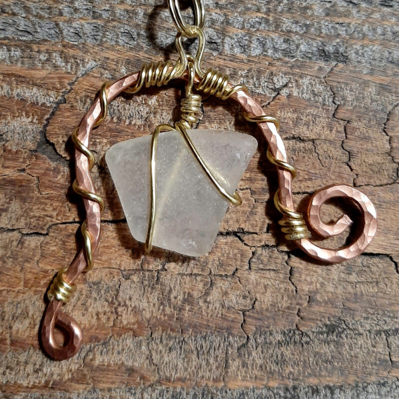 A piece of white creek glass is suspended between the arms of a rough arch of hammered copper wire, both wound round with much thinner golf-colored wire. One end of the arch ends in a spiral; the other extends further downward, and ends in a loop.