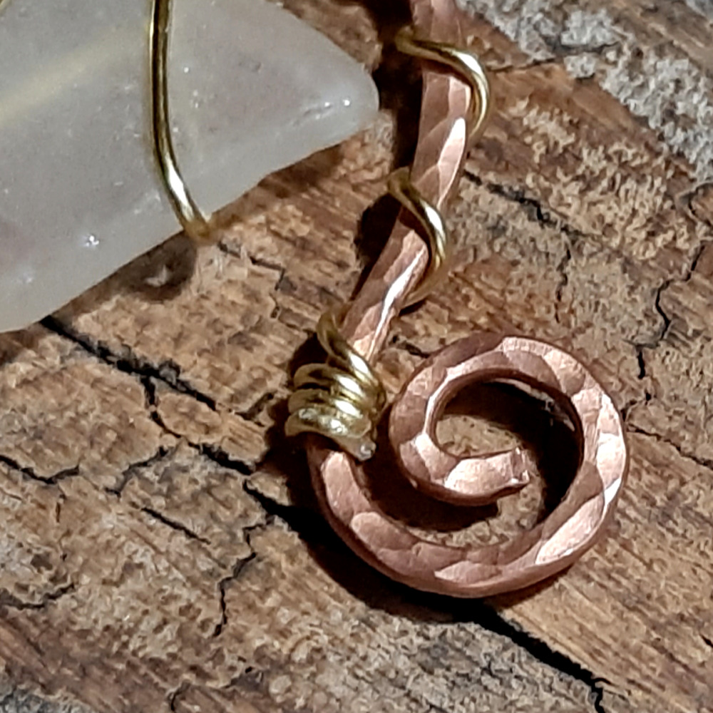A piece of white creek glass is suspended between the arms of a rough arch of hammered copper wire, both wound round with much thinner golf-colored wire. One end of the arch ends in a spiral; the other extends further downward, and ends in a loop.