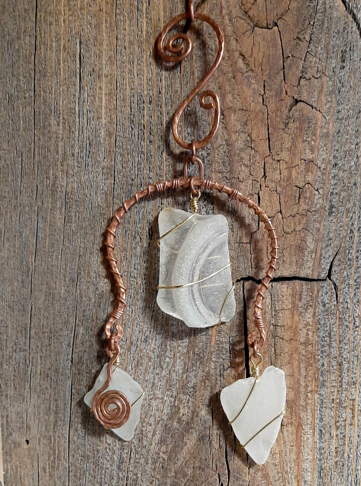 This pixy home decor piece features a broad arch of hammered copper wire wrapped round with thinner copper wire. Between the arms of the arch is a piece of white creek glass simply wrapped in gold-colored wire; smaller pieces hang from the bottom of each arm. A hammered copper spiral hangs in front of the left one.
