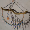 This faery home decor piece has an interestingly bumpy piece of thin driftwood, suspended horizontally by three chains whose links are decorated with blue beads, all of which hang from a hammered copper hook. From the driftwood hangs a copper spiral and two arcs of hammered copper, all three decorated with blue and green beads.