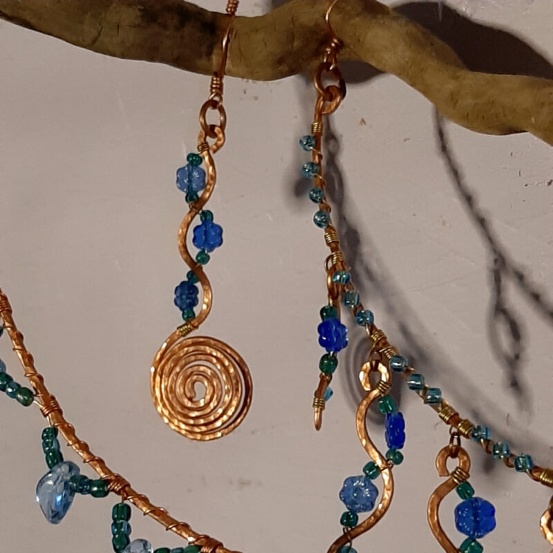 This faery home decor piece has an interestingly bumpy piece of thin driftwood, suspended horizontally by three chains whose links are decorated with blue beads, all of which hang from a hammered copper hook. From the driftwood hangs a copper spiral and two arcs of hammered copper, all three decorated with blue and green beads.