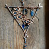 This faerie home decor piece features a point-down triangle of heavy hammered aluminum wire. The hook at the top is the same wire; below it hangs a hammered copper spiral. Much of the triangle is taken up by meandering copper wire; two blue glass leaves hang therein, along with a brass circle, and glass pony beads in green and blue.