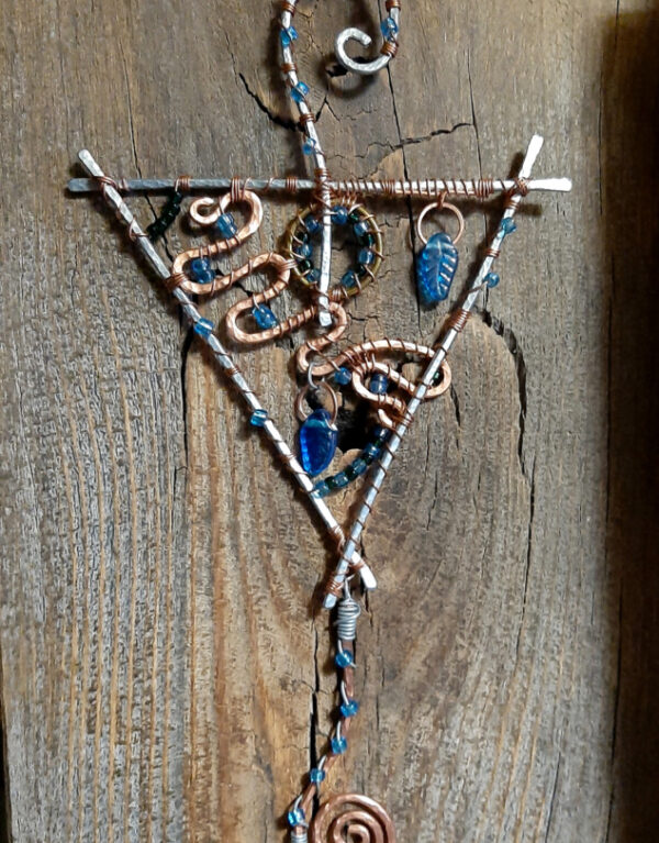 This faerie home decor piece features a point-down triangle of heavy hammered aluminum wire. The hook at the top is the same wire; below it hangs a hammered copper spiral. Much of the triangle is taken up by meandering copper wire; two blue glass leaves hang therein, along with a brass circle, and glass pony beads in green and blue.