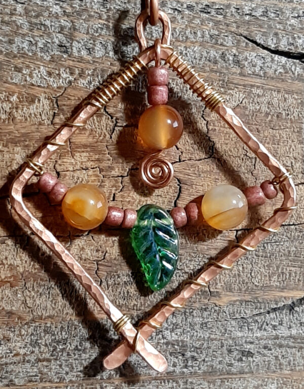 A rectangle of hammered copper wire, turned 45 degrees and crossed by a green glass leaf bead strung on fine copper along with brown and striped orange beads.