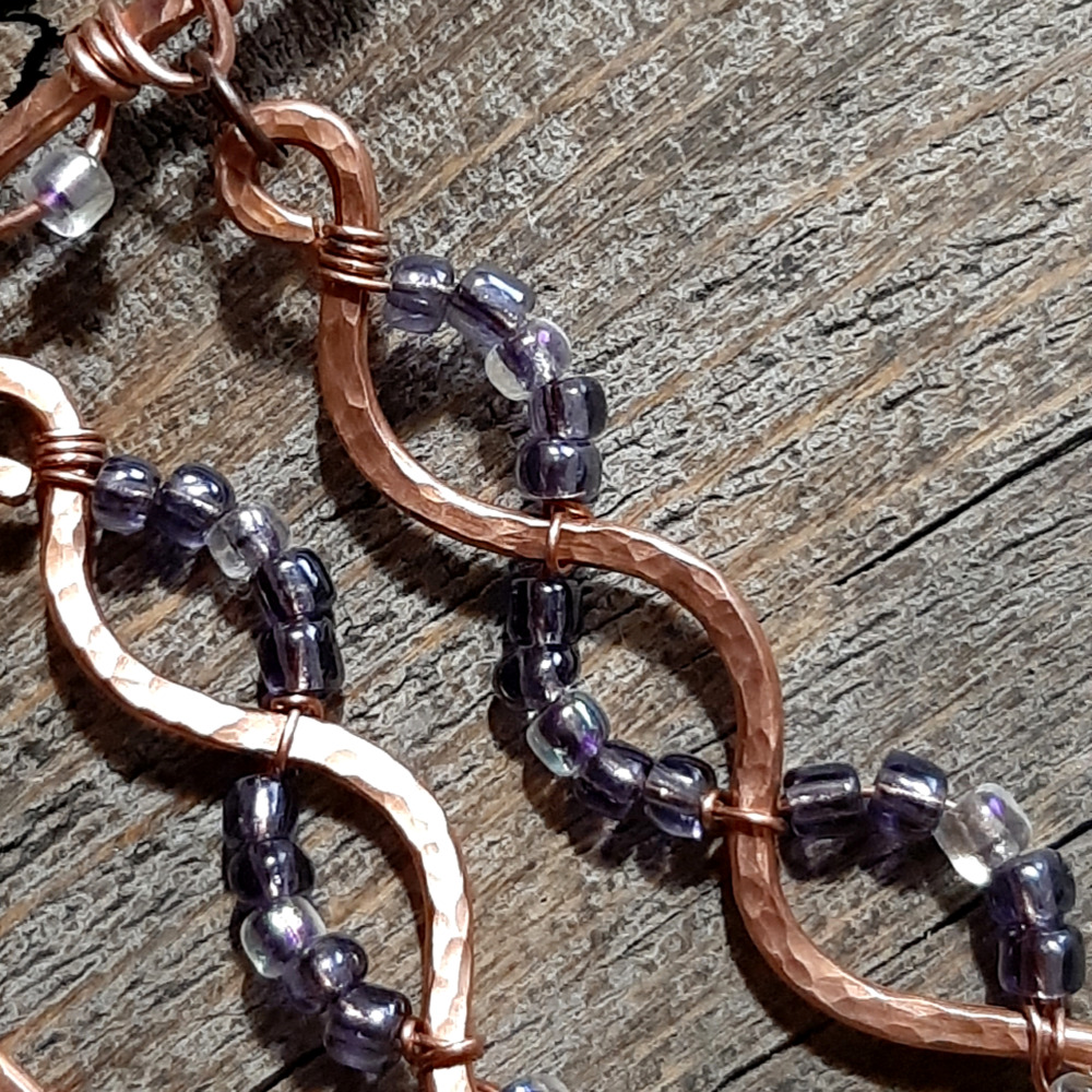 A broad horizontal bar of hammered copper wire, from which hang five meandering lengths of the same copper, each with purple and clear glass beads wired to them in complementary arcs.
