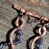 A broad horizontal bar of hammered copper wire, from which hang five meandering lengths of the same copper, each with purple and clear glass beads wired to them in complementary arcs.