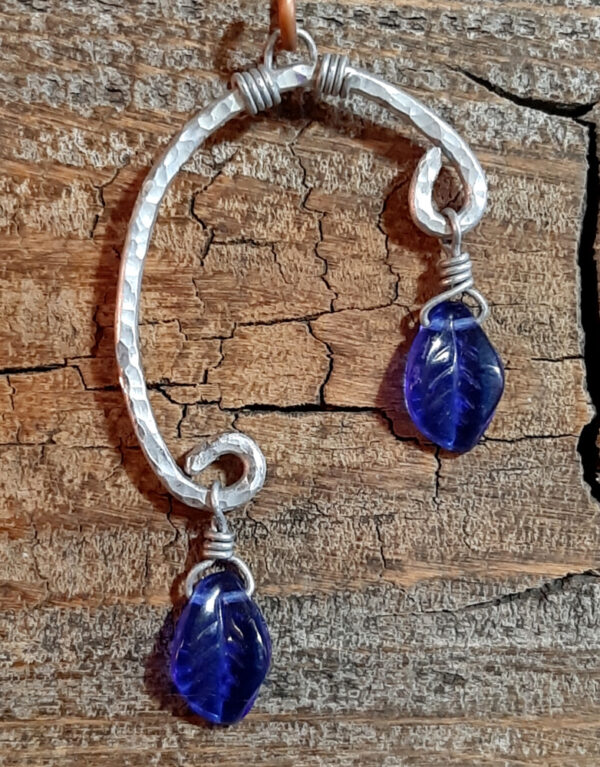 An uneven arc of hammered aluminum wire, with one end much higher than the other. From both ends hang cobalt blue glass leaves.