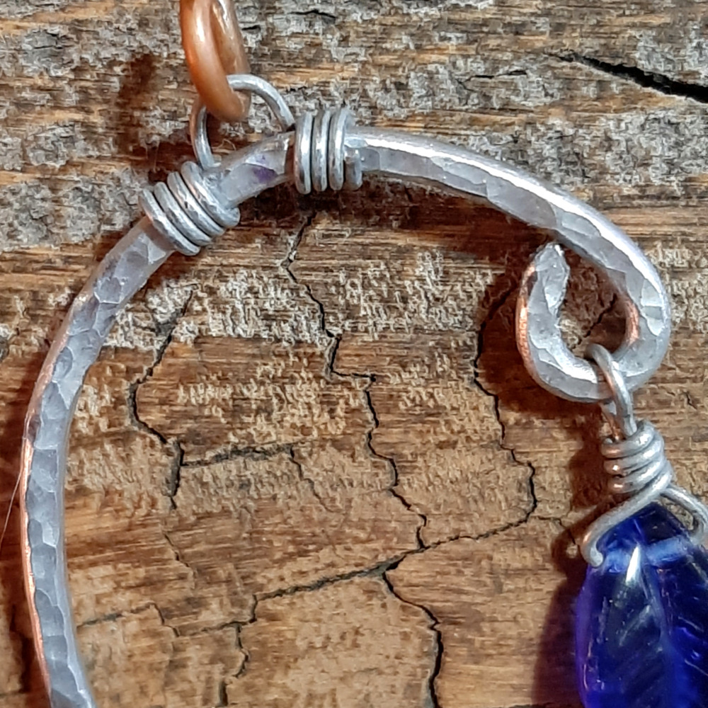 An uneven arc of hammered aluminum wire, with one end much higher than the other. From both ends hang cobalt blue glass leaves.