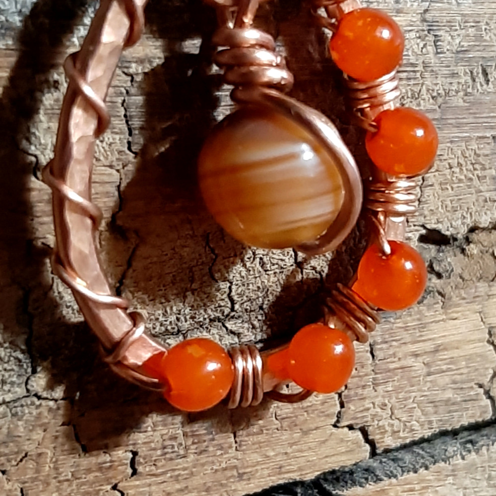 A simple teardrop shape in hammered copper wire, with round orange glass beads fastened around in thinner copper. A striped brown and white glass bead hangs in the center.