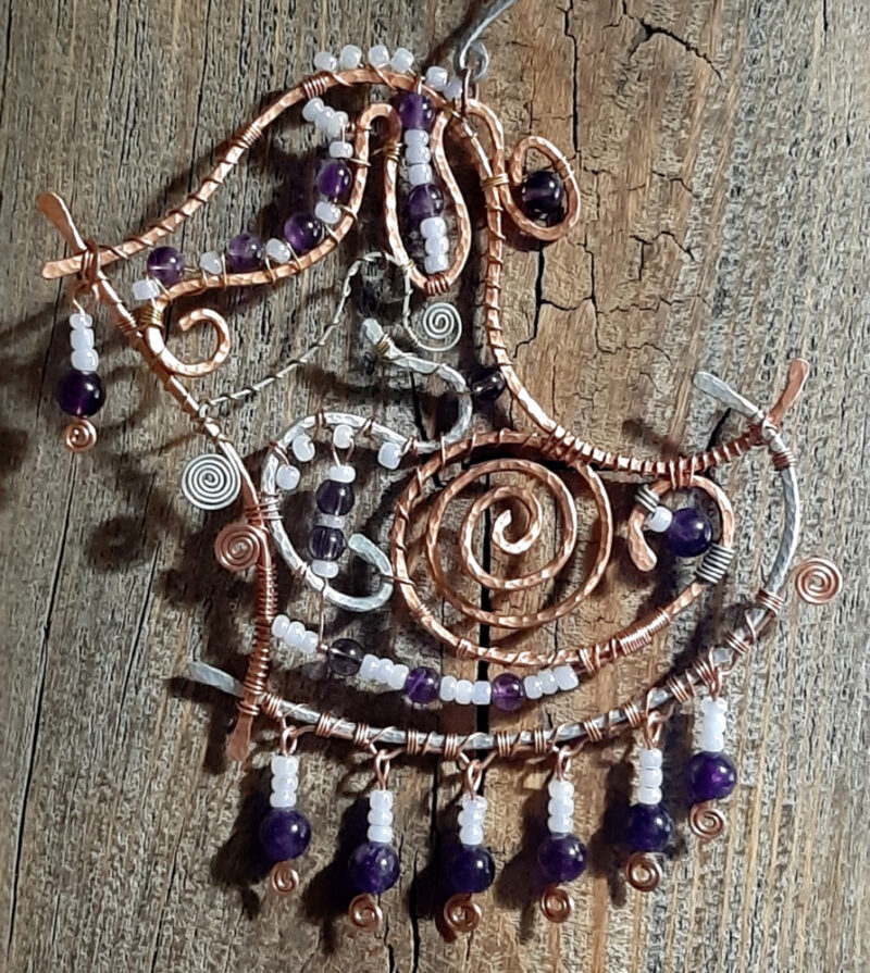 This faerie home decor piece is irregularly shaped, bounded by curved lengths of hammered copper and aluminum wire. The space within is festooned with more wire in tighter curves. Amethyst and white glass beads are strung throughout.