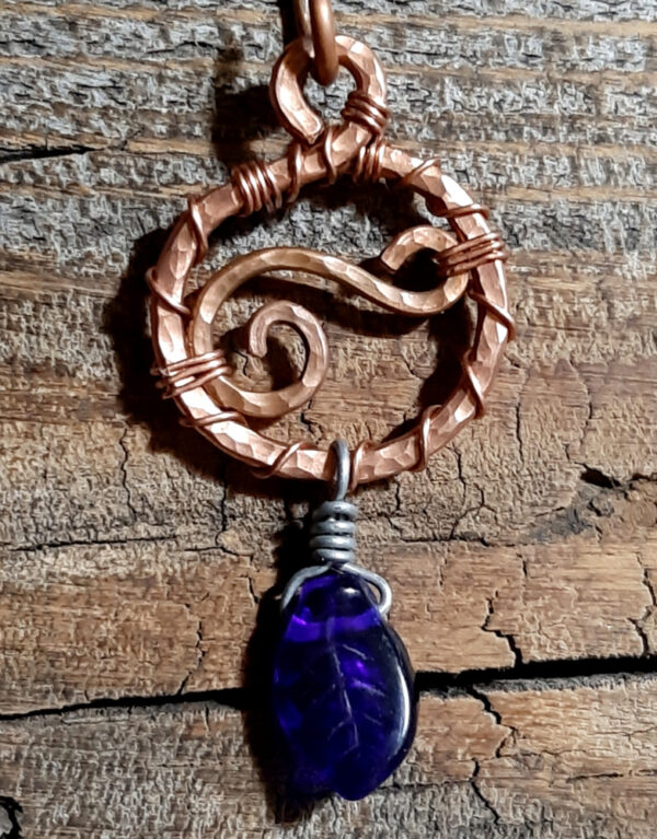This pendant features a cobalt blue glass leaf hanging from a circle of hammered copper wire, which has, also, a stylized S fastened within it horizontally.
