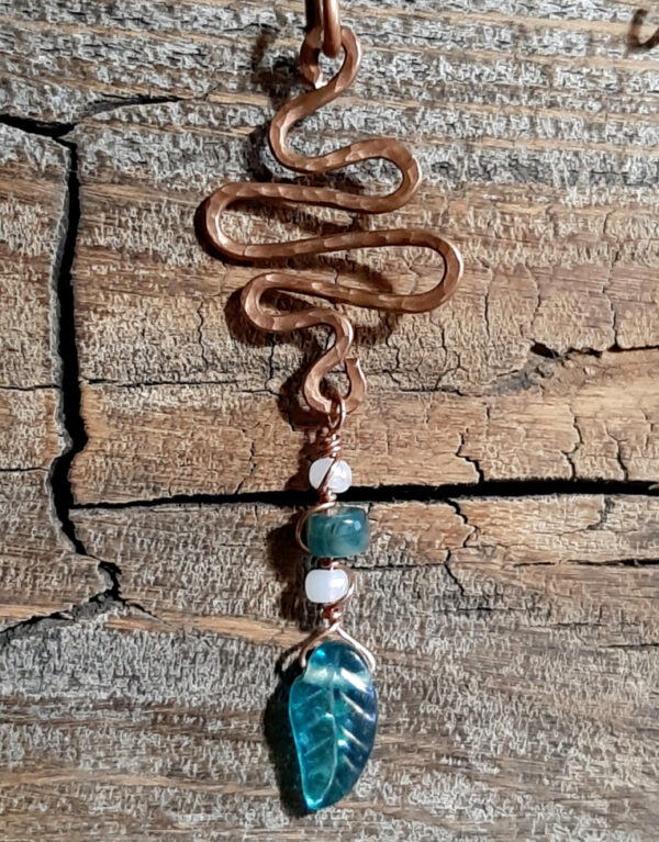 A squiggle of hammered copper wire with loops at top and bottom; from the bottom hangs a green glass leaf, strung on finer copper wire along with white and green beads.