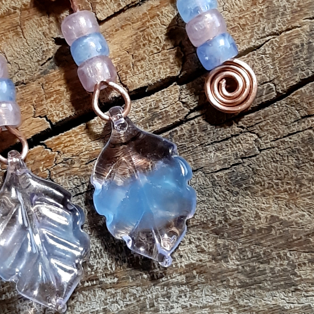 Each earring is an uneven arch of hammered copper, with three pink and pale blue beads hanging from the short end, and three pink and pale blue beads with a big clear, pink, and blue leaf hanging from the other.